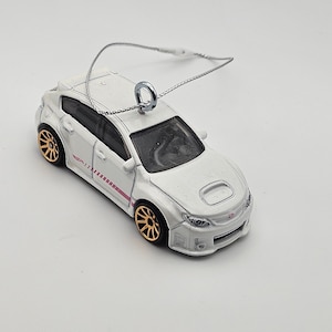 May include: A white Subaru Impreza WRX STI model car ornament with gold wheels and a pink stripe. The car is hanging from a silver string.