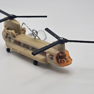 May include: A tan-coloured military transport helicopter ornament featuring two black rotor blades, an orange translucent cockpit window, and a silver hanging loop. The fuselage displays the number 72 and a small emblem on the side.