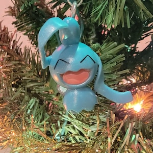 May include: A blue and pink Christmas ornament shaped like a cartoon character with a wide smile. The ornament is hanging from a silver string on a green Christmas tree.