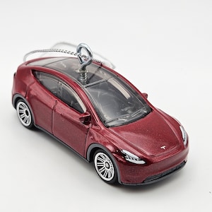 May include: A red Tesla Model Y ornament with a silver hook for hanging. The ornament is made of plastic and has a clear plastic roof.