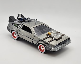 May include: Silver DeLorean toy car with red and white wheels, and detailed engine components. The car has a brushed metal finish and is a replica of the iconic vehicle from the Back to the Future movie series. A silver loop is attached for hanging.