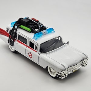 May include: White toy car with a red and white stripe, a ladder on the side, and a green and black contraption on the roof. The car has a blue light on the roof and a ghost logo on the side.