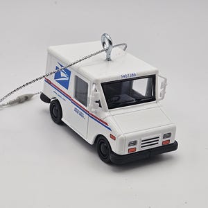 May include: A white USPS mail truck ornament with black wheels and red and blue stripes. The ornament has a silver hanging loop and a silver chain. The text "www.usps.com" is visible on the side.