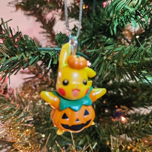 May include: A yellow Pikachu Pokemon character ornament dressed as a jack-o'-lantern with a green stem and orange pumpkin face. The ornament is hanging on a green Christmas tree.
