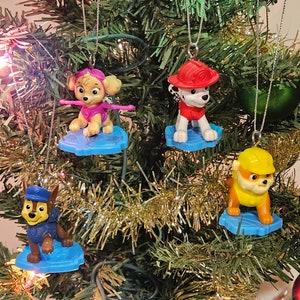 May include: Four Paw Patrol characters on blue snowboards, decorated as Christmas ornaments. The characters are Chase, Skye, Marshall, and Rubble.