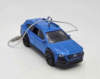 May include: A bright blue die-cast toy car ornament featuring black wheel arches, black wheels, and a silver metal eyelet screw attached to the roof. A thin silver cord is looped through the eyelet for hanging, with a small grey plastic fastener.