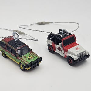May include: Two miniature toy cars, one a green and yellow SUV with "Jurassic Park" branding, the other a red and white Jeep with "JP18" on the hood. Both have silver loops for hanging.