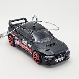 May include: Black die-cast model car with red wheels and accents. The car has a spoiler and the text "368 gravel crew". A silver chain is attached to the top of the car for hanging.