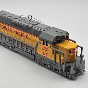May include: A yellow and grey model train featuring the words "POWER PACIFIC". The number "65" is displayed on the front. The train has a silver hanging loop for display.