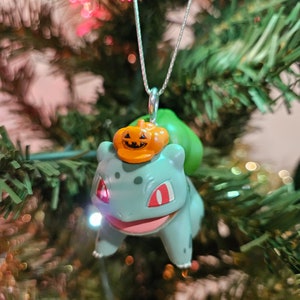 May include: A green and blue Pokemon bauble with a pumpkin hat hanging on a Christmas tree. The bauble is a Bulbasaur, a grass-type Pokemon.