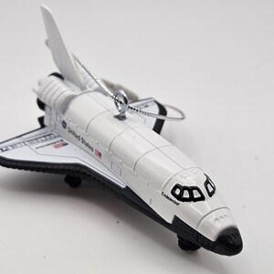 May include: A white and black space shuttle ornament with the text "Endeavour" on the front. The ornament has a silver string for hanging and features the United States flag. The wings and tail are black.
