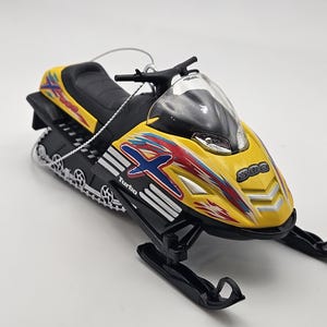 May include: Yellow and black snowmobile ornament with red and blue accents. The snowmobile has a clear windshield and the text "Turbo" and "300" on the side.