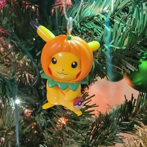 May include: A yellow Pikachu Pokemon character wearing a pumpkin costume with a green leaf collar and a red Pokeball on its side. The character is hanging on a green Christmas tree.