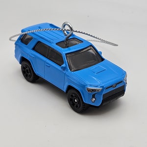 May include: A die-cast, light blue SUV toy with black wheels and a silver chain for hanging. The miniature vehicle features detailed headlights, a black grille, and a sunroof. A small Toyota logo is visible on the front.