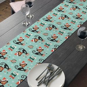 May include: A turquoise table runner featuring a repeating pattern of black cats wearing Santa hats and holding gifts. The design includes snowflakes and retro-style Christmas elements. The runner is displayed on a dark wooden table, with a white plate and silverware.
