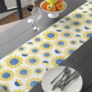 May include: A white table runner with a yellow and blue sunflower pattern. The runner is laid on a dark wood table with a plate and silverware set on top.