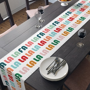 Op de afbeelding: Een witte tafelrunner met een herhalend patroon van "la la la" in kleurrijke, afgeronde, geometrische vormen. De runner ligt op een donkerhouten tafel met twee wijnglazen en een bord met bestek.