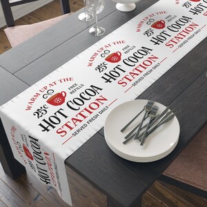 May include: White table runner with red text that reads "Warm up at the 25¢ Hot Cocoa Station - Served Fresh Daily". The runner is on a dark wood table with a white plate and silverware.