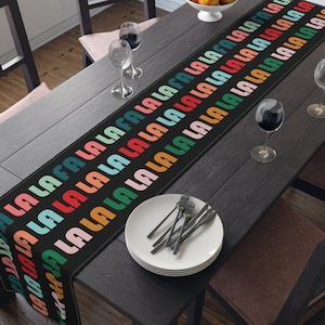 Op de afbeelding: Een zwarte tafelrunner met een herhalend patroon van het woord "la la" in regenboogkleuren. De runner ligt op een zwarte tafel met twee wijnglazen en een kom met fruit.