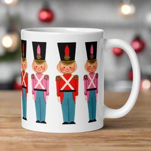 May include: White ceramic mug with a design of five toy soldiers in a row. The soldiers are wearing red, pink, and blue uniforms with black hats and white trim.