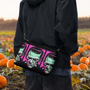 May include: A black crossbody bag with a pink and black print of Frankenstein's face. The bag is worn by a person standing in a field of pumpkins.