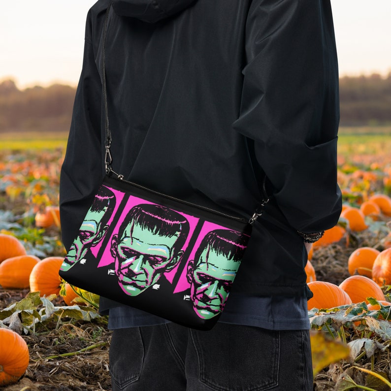 May include: A black crossbody bag with a pink and black print of Frankenstein's face. The bag is worn by a person standing in a field of pumpkins.