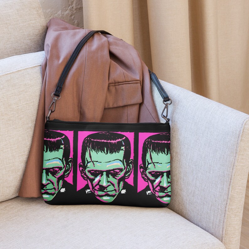 This black crossbody shoulder bag features a retro gothic print of Frankenstein's Monster with a green face and pink background. It's printed in a pattern on both sides.