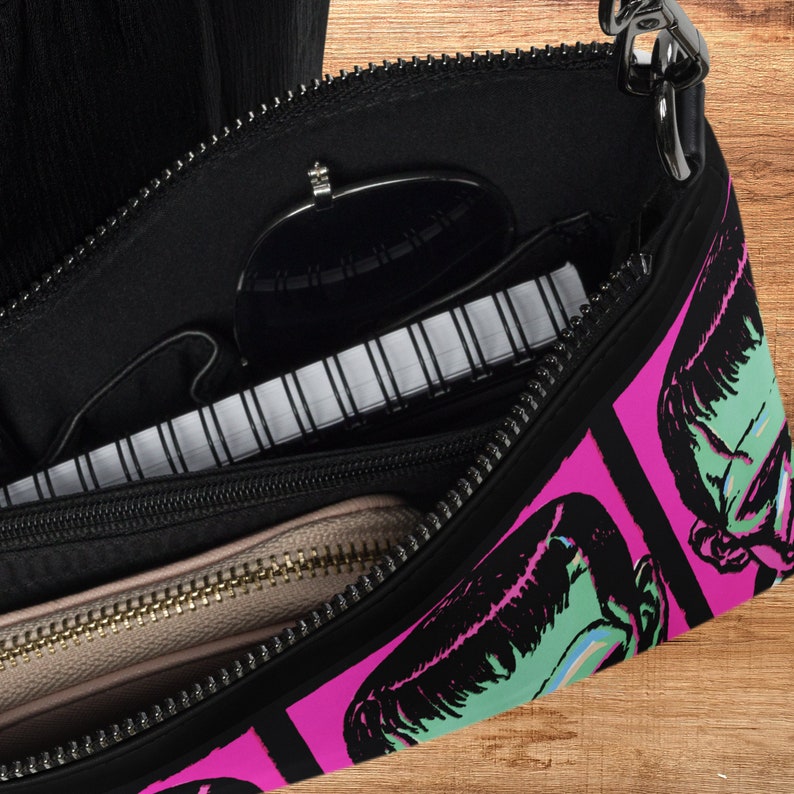 This shoulder crossbody purse converts to a large wristlet clutch. This purse features a retro Frankenstein Monster print of a green monster with a pink background.