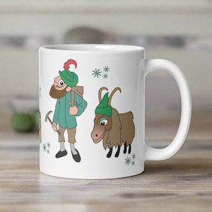May include: White ceramic mug with a green and brown illustration of a man with a pickaxe and a goat. The man is wearing a green hat with a red feather. The background has a snowflake pattern.