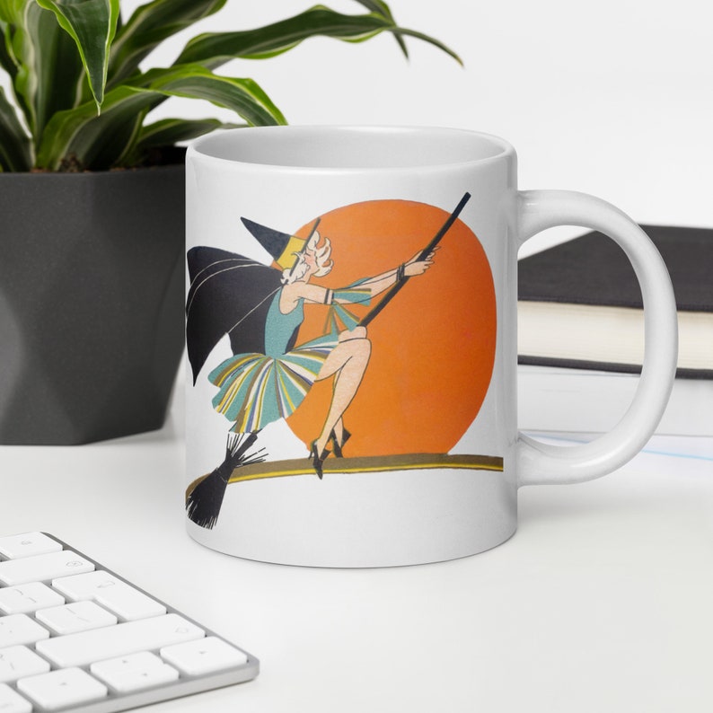 This ceramic coffee mug features a vintage deco illustration of a cute flapper witch flying on a broom with an orange full moon behind her.