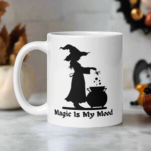 May include: White ceramic mug featuring a black silhouette of a witch stirring a cauldron. The text "Magic Is My Mood" is printed on the mug.
