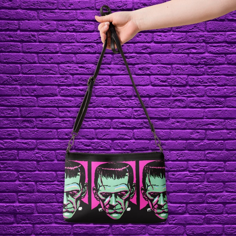 This black crossbody shoulder bag features a retro gothic print of Frankenstein's Monster with a green face and pink background. It's printed in a pattern on both sides.