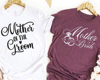 May include: Two t-shirts, one white and one burgundy, with wedding-themed text. The white shirt says "Mother of the Groom" in black script. The burgundy shirt says "Mother of the Bride" with a ring graphic. Gold hangers and floral accents.