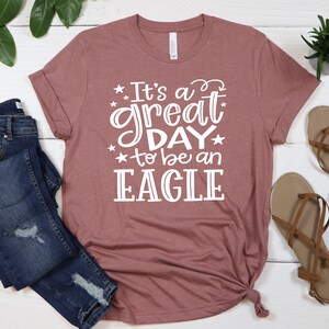 K&ouml;nnte beinhalten: Ein mauvefarbenes T-Shirt mit dem Text "It's a great day to be an EAGLE" in wei&szlig;en Buchstaben.