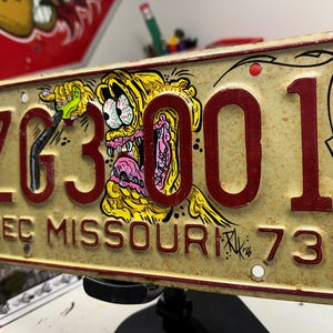 Puede incluir: Una matrícula vintage de Missouri de diciembre de 1973 con un acabado crema desgastado y caracteres en relieve de color rojo oscuro que dicen ZG3001. La placa presenta un personaje de dibujos animados amarillo pintado a mano.