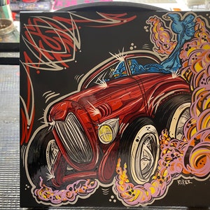May include: A square, black artwork featuring a red hot rod car with flames and a figure in the driver's seat. The car has white-walled tires and is set against a black background with red and white pinstriping. The artwork is signed "RLK22".