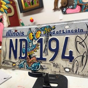 May include: An Illinois license plate with the text "ND 194" in blue. The plate is decorated with a cartoonish blue and yellow creature. The words "Land of Lincoln" are at the top. Black abstract art is on the right side.