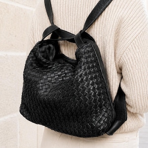 Pode incluir: Mochila de couro preto tecido com alças ajustáveis. A bolsa tem um design macio e folgado e um padrão tecido. As alças são pretas e parecem ser de couro. A mochila é usada sobre os ombros.