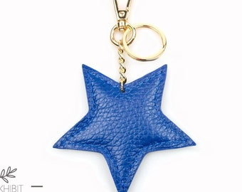 May include: A star-shaped keychain crafted from textured blue leather, featuring a gold-toned metal lobster clasp and a circular split ring attached by a short, twisted gold-toned chain. The design is simple, with visible stitching along the edges.