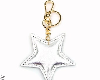 May include: Silver leather star keychain with a gold chain and clasp. The star has a stitched design and is attached to a gold ring.