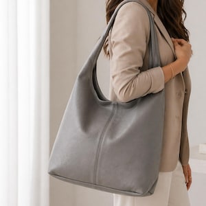 May include: A large, grey leather hobo bag with a curved handle. The bag has a simple, minimalist design with a vertical seam down the centre. The bag is carried over the shoulder.