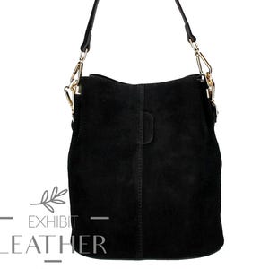 Puede incluir: Bolso de hombro de ante negro con herrajes dorados y una correa larga y ajustable. El bolso tiene forma rectangular con la parte superior curvada y un asa pequeña. El texto "EXHIBIT LEATHER" está impreso en la parte inferior.