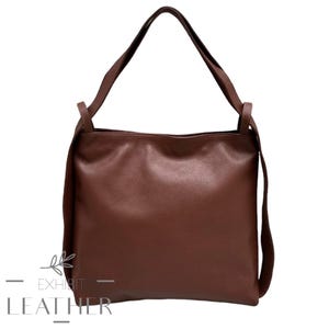 May include: A brown leather tote bag with long shoulder straps. The bag has a square shape and a textured surface. The text "EXHIBIT LEATHER" and "Welcome To A New Look" is visible at the bottom of the image.