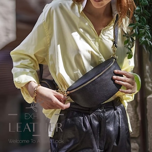 Puede incluir: Un bolso de cintura de cuero negro con cremallera y herrajes dorados. El bolso se lleva cruzado. La imagen también muestra una pulsera dorada y el texto "EXHIBIT LEATHER" y "Welcome To A New Look".