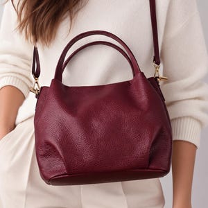 May include: A deep burgundy handbag with a textured surface, featuring two rounded top handles and a detachable shoulder strap with gold-toned metal hardware. The bag has a structured base and a soft, slouchy body design with visible stitching.