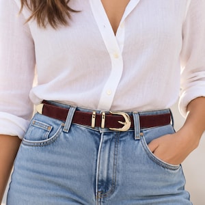 Op de afbeelding: Een donkerbordeaux suède riem met een gepolijste goudkleurige metalen gesp en een bijpassende metalen punt. De riem wordt gedragen met een lichtblauwe spijkerbroek en een wit overhemd, wat zorgt voor een klassieke en strakke uitstraling.