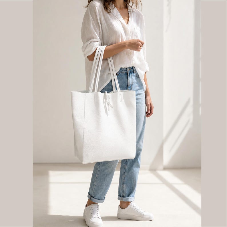 Pode incluir: Uma bolsa tote grande de cor branca s&oacute;lida com uma superf&iacute;cie lisa e estruturada, apresentando duas al&ccedil;as longas de ombro. A bolsa possui um detalhe de costura vertical simples no painel frontal e um pequeno fecho de la&ccedil;o centralizado no topo.