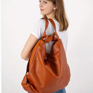 Peut inclure: Un grand sac hobo en cuir couleur cognac doté d'une silhouette souple et déstructurée, de deux anses supérieures et d'un compartiment principal spacieux. Le sac est porté à l'épaule, mettant en valeur sa texture lisse et son ton brun chaud.