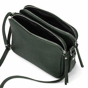 May include: A dark green crossbody bag with three zipped compartments. The bag has a long, adjustable strap and silver-toned hardware. The interior of the bag is black. The bag is made of a textured material.