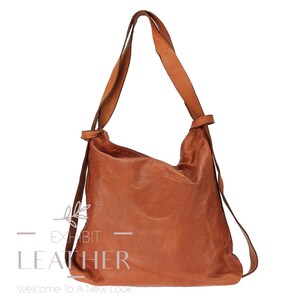 May include: A brown leather shoulder bag with a long strap and a smaller strap that can be used as a backpack strap. The bag has the text "Exhibit Leather" printed on the front.